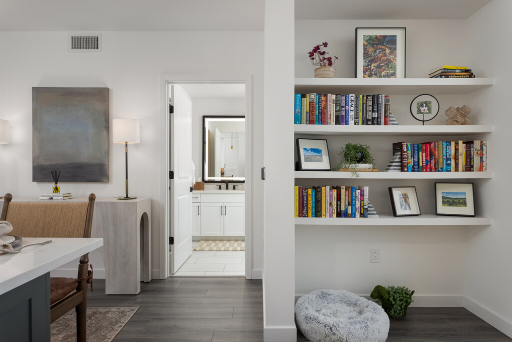 Contemporary home interior with built-in shelving displaying books and decor, open doorway to bathroom, kitchen island, and abstract artwork on white walls.