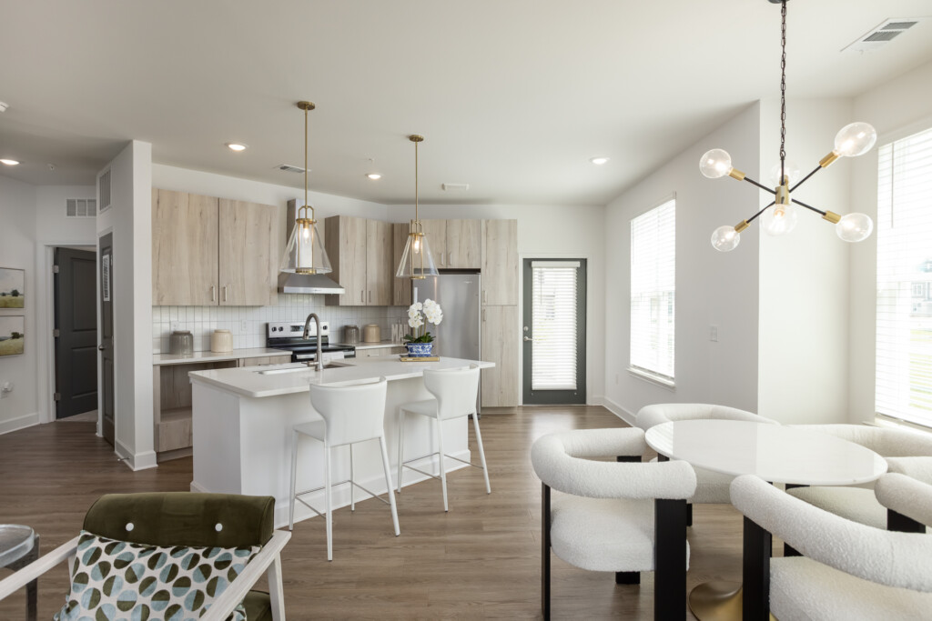 Open-concept kitchen and living area with light wood cabinetry, white island with seating, pendant lighting, curved cream-colored chairs, and bright windows