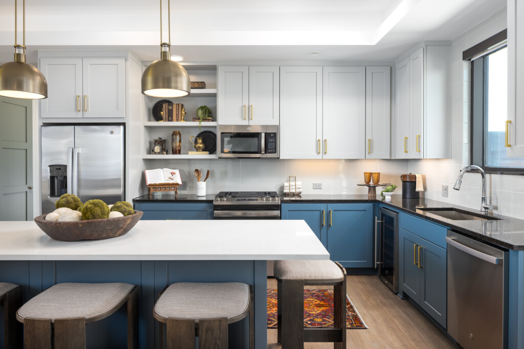 Contemporary kitchen featuring blue lower cabinets, white upper cabinets, stainless steel appliances, brass pendant lights, and white quartz island with seating.