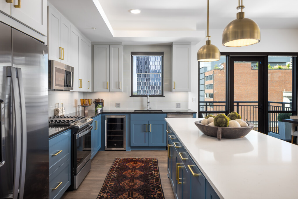 Contemporary kitchen with white upper cabinets, blue lower cabinets, stainless steel appliances, white countertops, brass pendant lights, and black-framed glass doors opening to a balcony.
