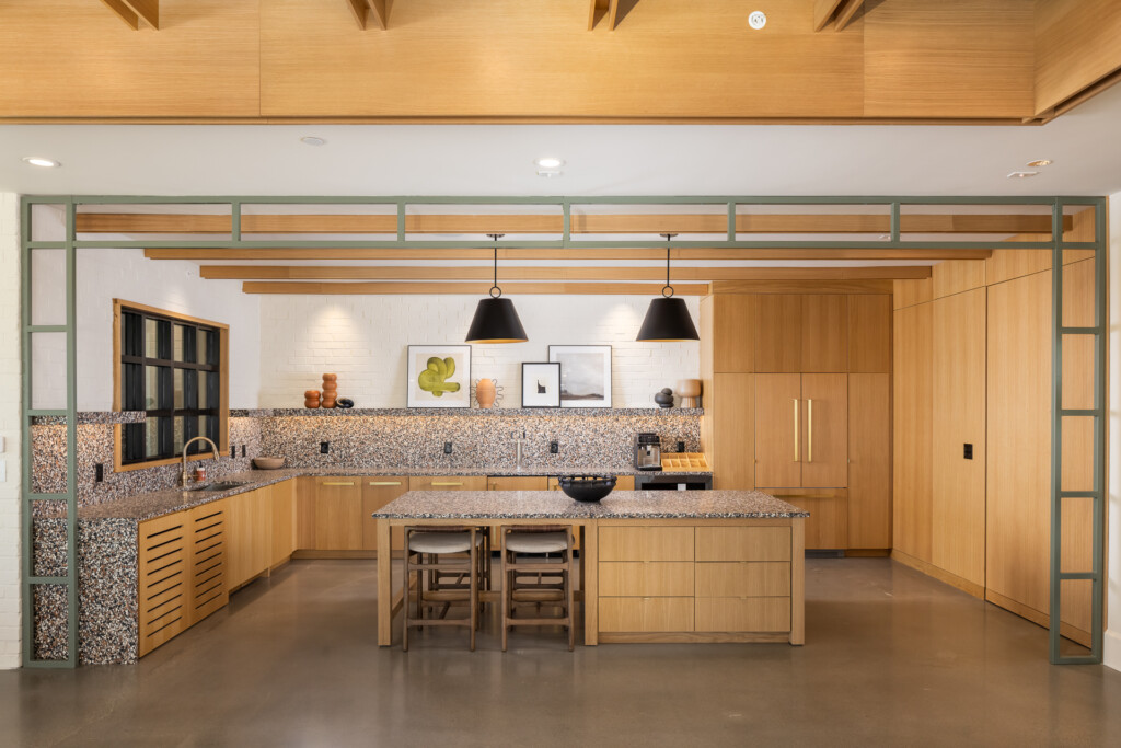 Contemporary kitchen with natural wood cabinetry, terrazzo backsplash, green-framed glass doors, central island with seating, and pendant lighting overhead.
