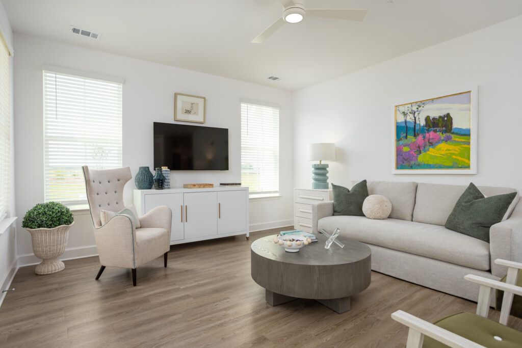 Bright living room with cream sofa, beige accent chair, oval wood coffee table, wall-mounted TV, and colorful landscape artwork on walls
