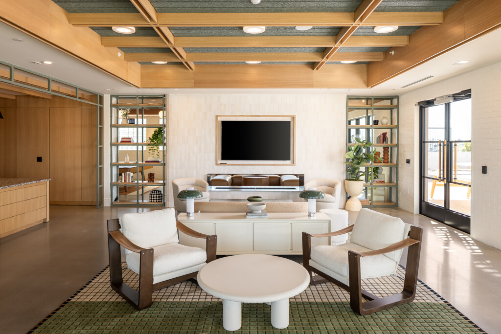 Bright living room with two cream armchairs facing a round white table, built-in shelving units, wood beam ceiling with recessed lighting, and glass doors leading to a terrace