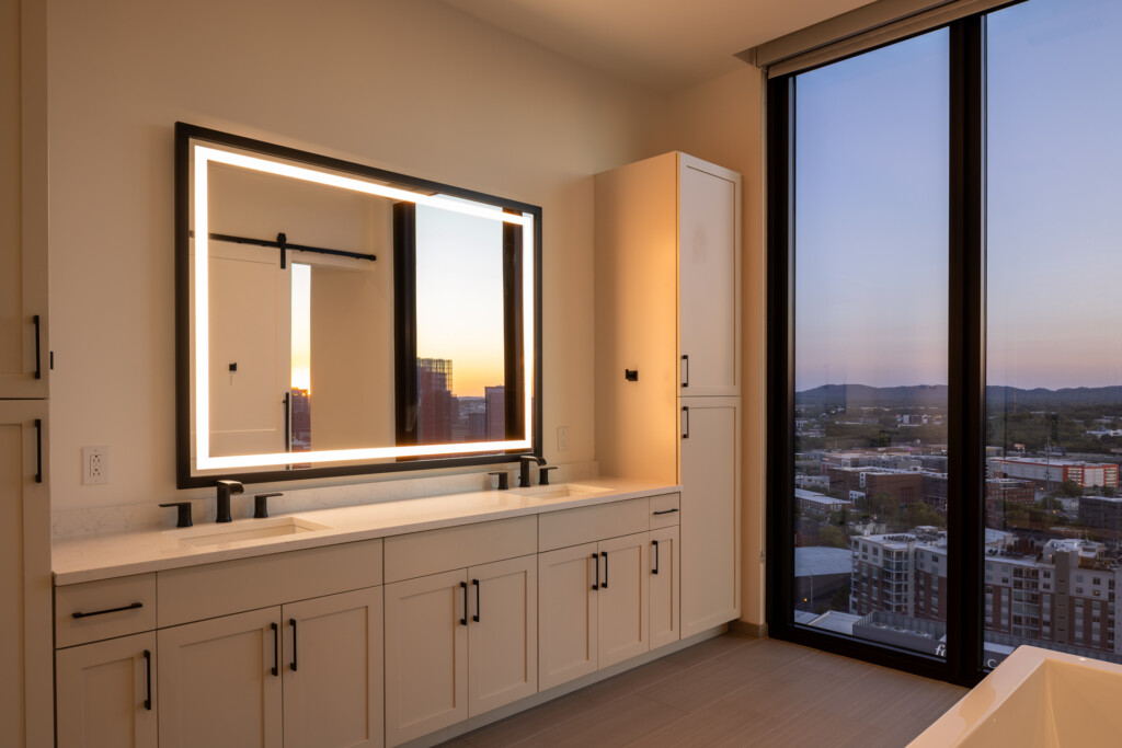 Spacious bathroom vanity with illuminated mirror, cream cabinetry, and floor-to-ceiling windows overlooking a cityscape at sunset
