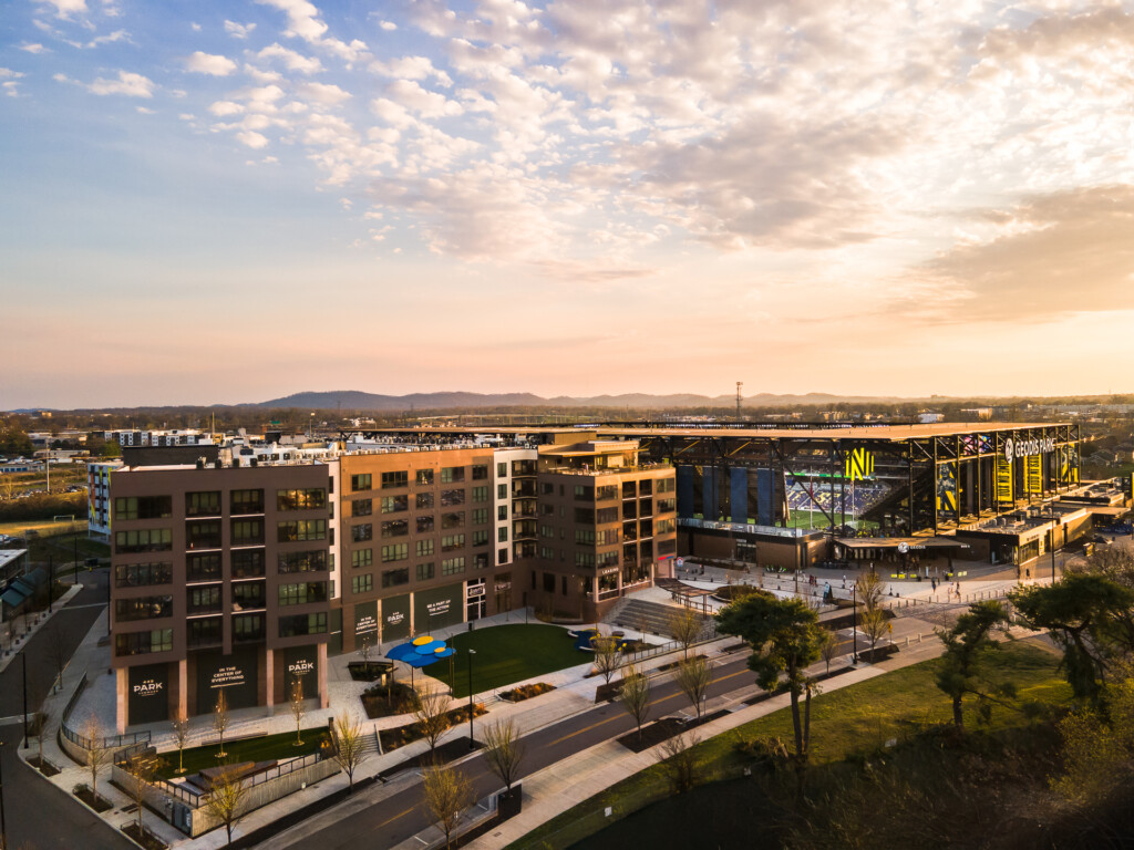 Mixed-use commercial and residential buildings with outdoor plaza, green space, and stadium in background at sunset