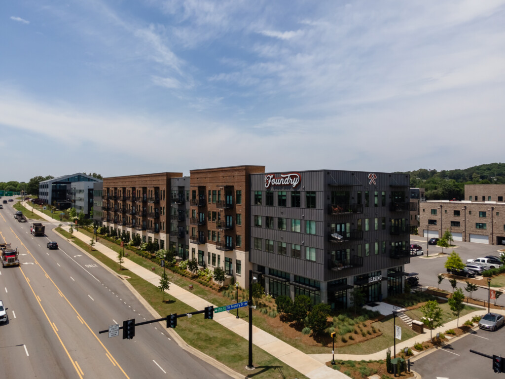 Contemporary multifamily residential and commercial buildings with brick and metal facades, ground-floor retail spaces, landscaped courtyards, and surface parking along a wide boulevard under blue skies.