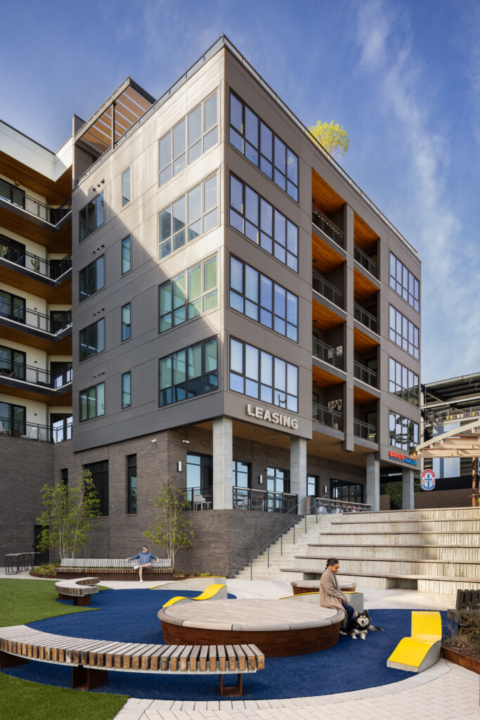 Contemporary residential building with large grid windows, concrete and brick facade, and ground-level courtyard featuring curved seating, blue surfacing, and landscaping.