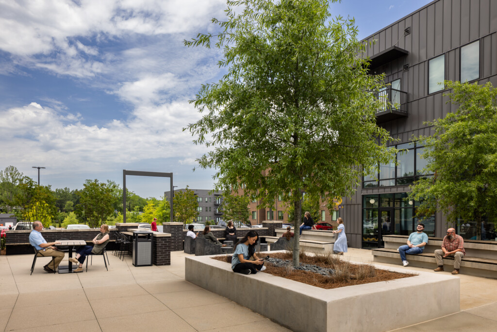 Open courtyard with concrete planter boxes, metal-frame seating, lush trees, and a contemporary dark-clad residential building with balconies overlooking the space.