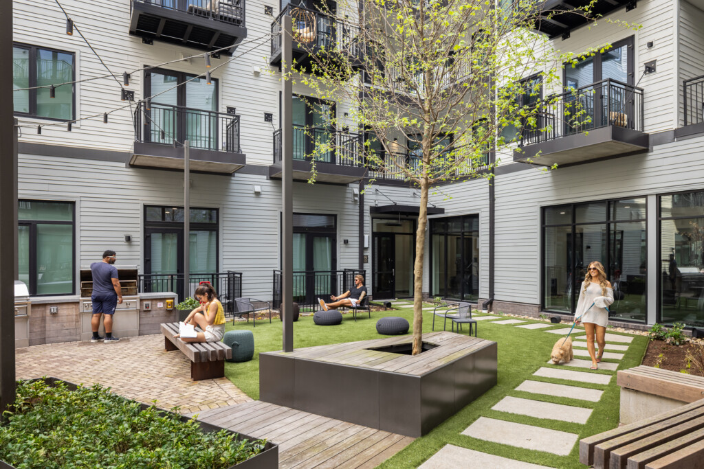 445 Park Commons - GEODIS - Contemporary apartment building courtyard with white siding, black railings, residents relaxing on benches and seating areas, central tree, manicured lawn, and paved walkways.