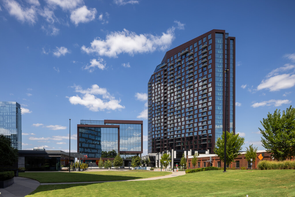 Tall residential building with brick and glass facade, contemporary mid-rise structures, and green lawn plaza under blue sky