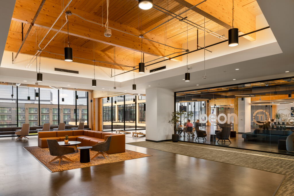 Open commercial lobby with exposed wood ceiling, pendant lighting, orange modern sofa, charcoal chairs, and glass-walled conference rooms