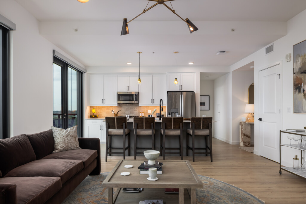 Olive at Peabody - Open-concept living space with white kitchen cabinetry, stainless steel appliances, island with seating, pendant lighting, brown leather sofa, and hardwood flooring throughout.