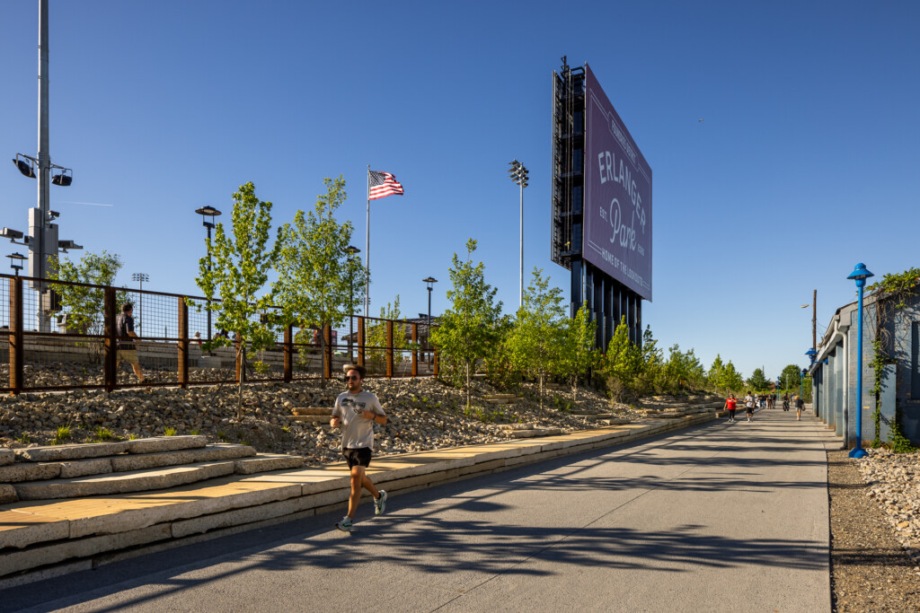 Runner jogging on a paved path through a contemporary public park with wooden log seating, landscaped hillside, American flag, and large digital display board under clear blue sky