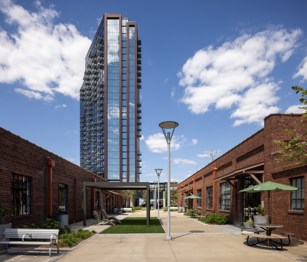 Olive at Peabody - Tall glass and metal residential tower rises behind a brick courtyard with seating areas, lamp posts, and green umbrellas on a sunny day