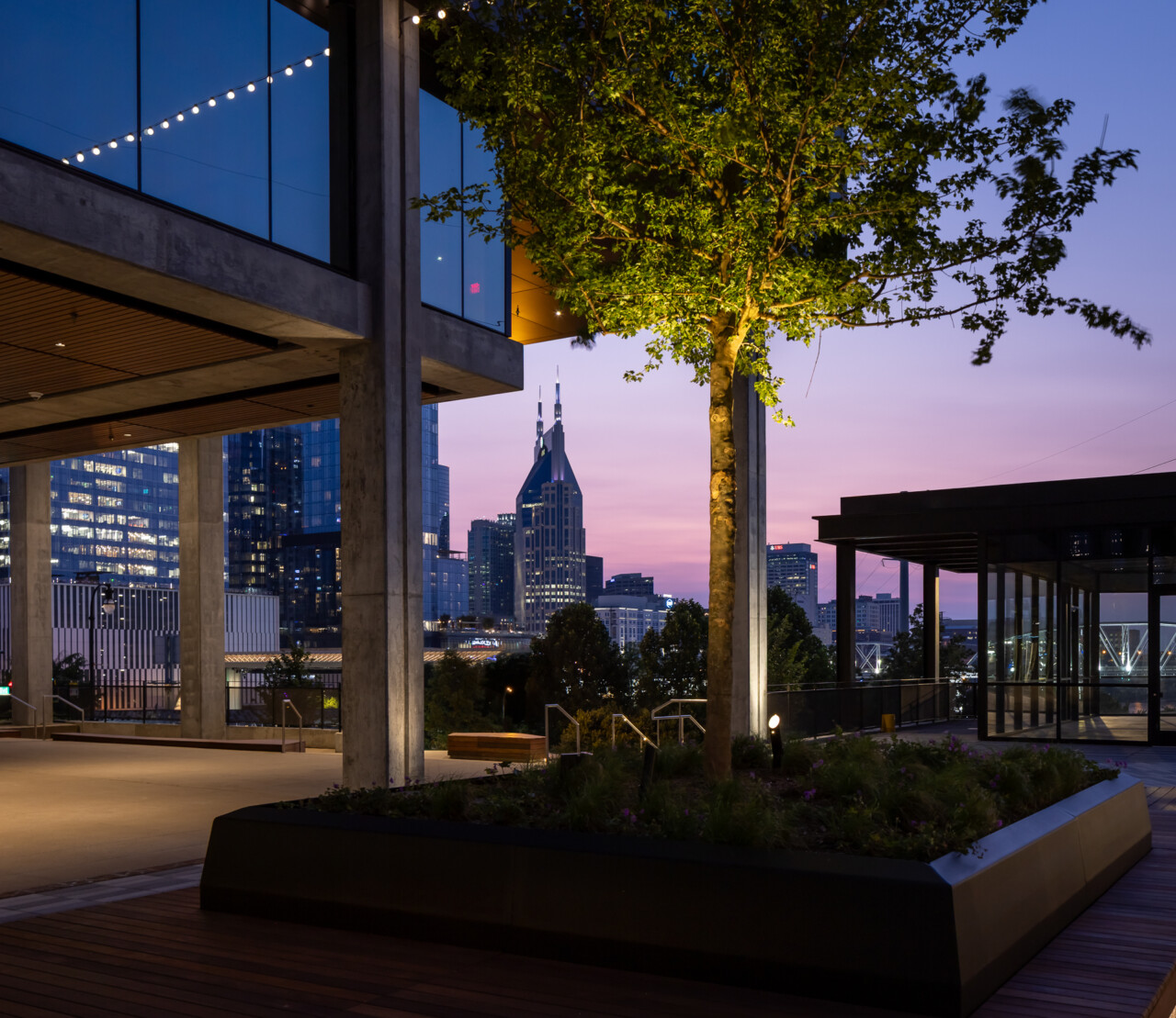 Olive at Peabody - Contemporary rooftop terrace with concrete columns, illuminated tree, planter beds, and floor-to-ceiling glass overlooking a downtown skyline at dusk