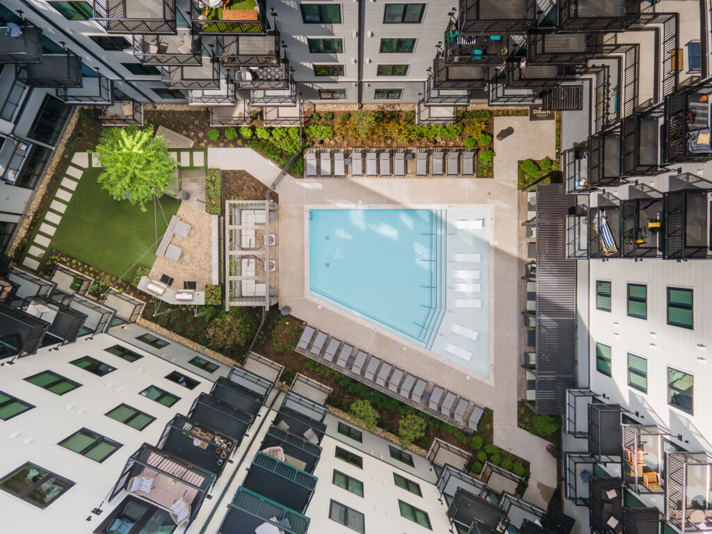 Aerial view of a modern multifamily residential complex featuring a central pool deck, surrounding apartments with balconies, landscaped green areas, and lounge seating.