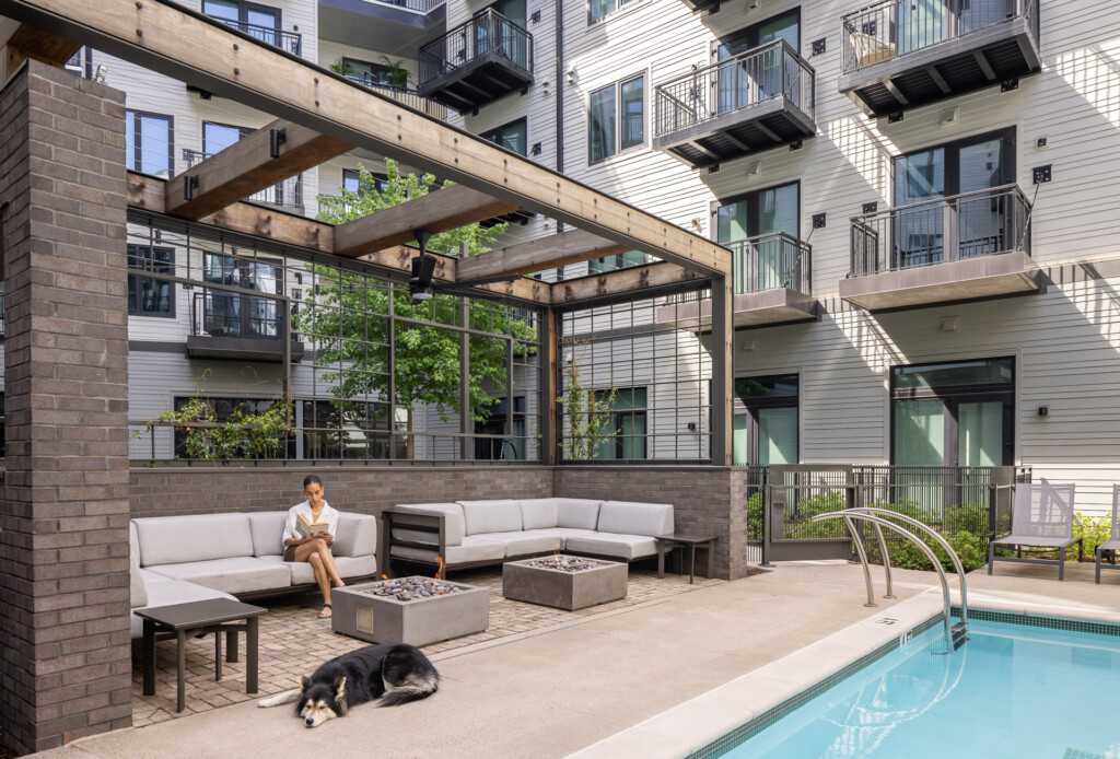 Residential courtyard with L-shaped cream seating, fire pits, wooden pergola, brick building, pool, and dog resting on pavers