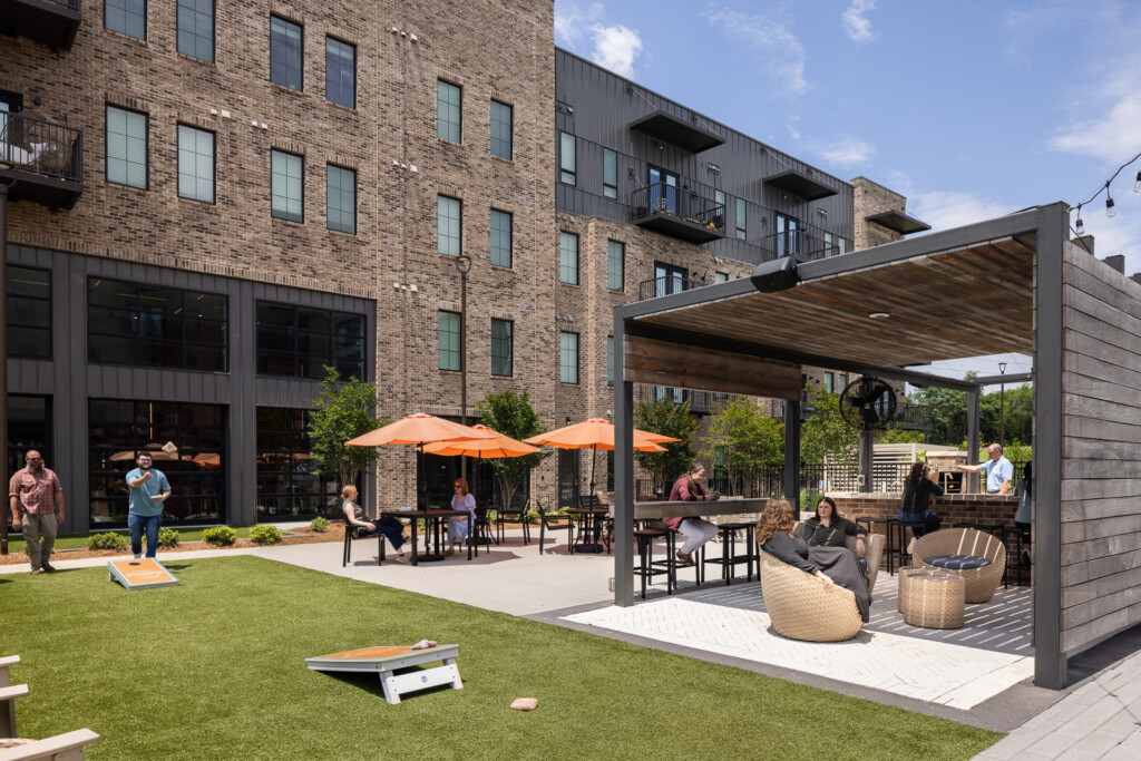 Modern courtyard with brick buildings, shaded seating areas under pergolas with orange umbrellas, green lawn, and residents relaxing on furniture and playing cornhole