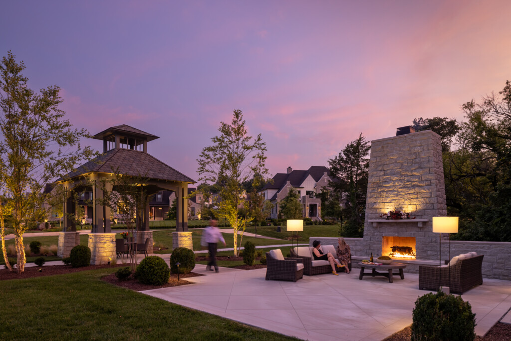 Illuminated backyard patio with outdoor fireplace, seating areas, gazebo structure, and residential homes visible beyond manicured landscaping at twilight.