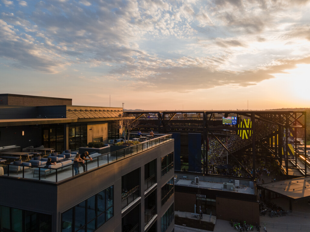 Rooftop terrace with seating areas overlooking a contemporary multifamily complex at sunset, featuring blue lounge furniture, steel railings, and dramatic cloudy sky.