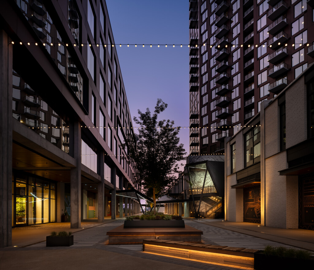 Olive at Peabody - Illuminated courtyard between tall residential buildings at twilight, with string lights overhead, wooden seating, and landscaping in the center