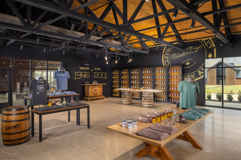 Clyde Mays Distillery - Industrial tasting room with black steel beam ceiling, golden skylights, bourbon barrel displays, retail shelving, and merchandise tables showcasing spirits and branded apparel.