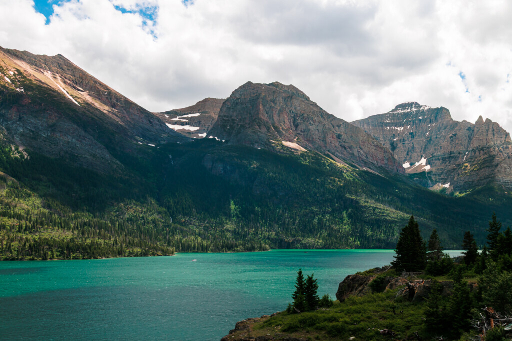 Turquoise alpine lake surrounded by evergreen forest with jagged mountain peaks and patches of snow above