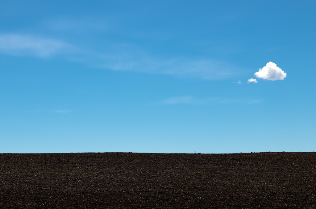 Minimalist landscape with a solitary white cloud against a bright blue sky above a dark soil horizon