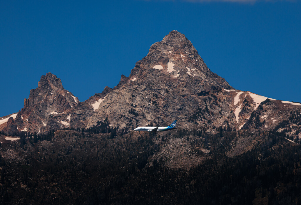 A commercial airplane with blue and white livery flies horizontally across a clear blue sky above a jagged mountain peak with snow patches and forested slopes below.