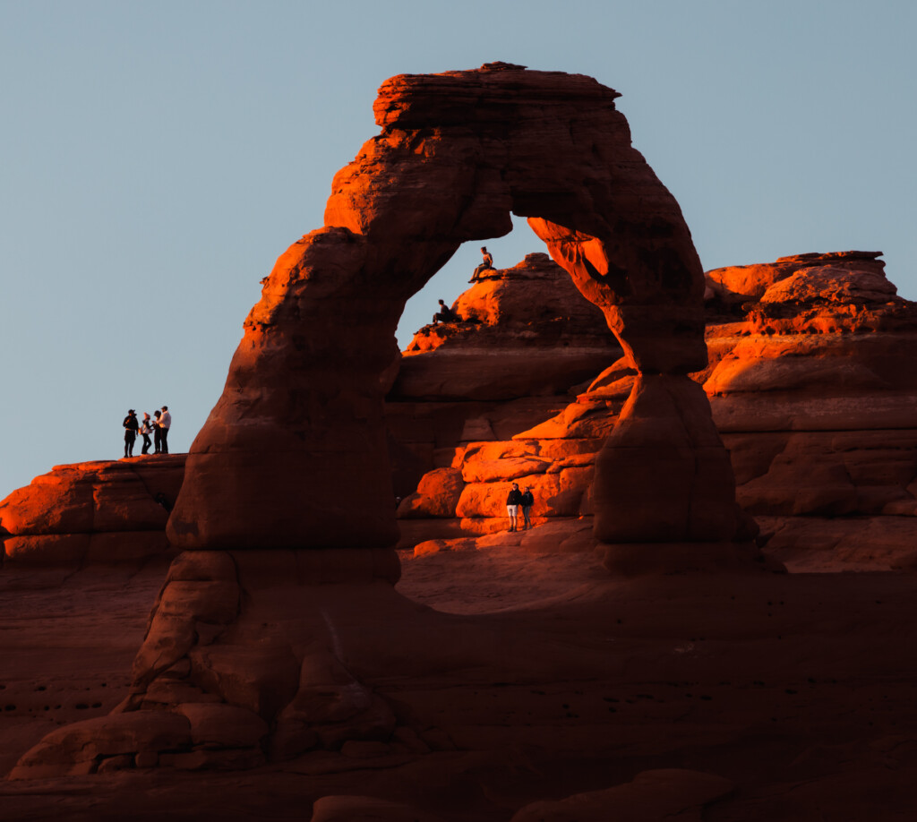 Red sandstone arch formation glowing orange in sunset light, with visitors scattered on the rock formations below