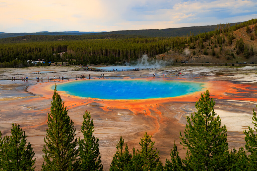Brilliant blue-green hot spring surrounded by orange and yellow mineral deposits, with evergreen forest and mountains beyond