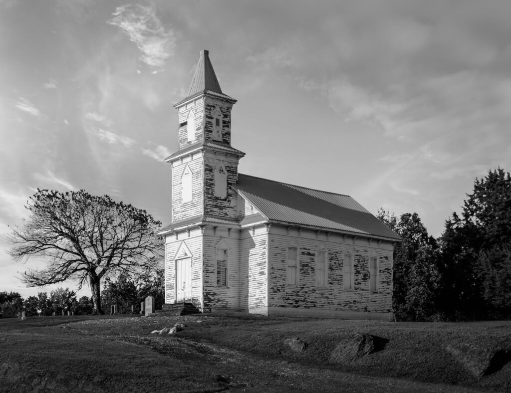 White wooden church with tall steeple and bell tower, metal roof, surrounded by bare and evergreen trees under cloudy sky
