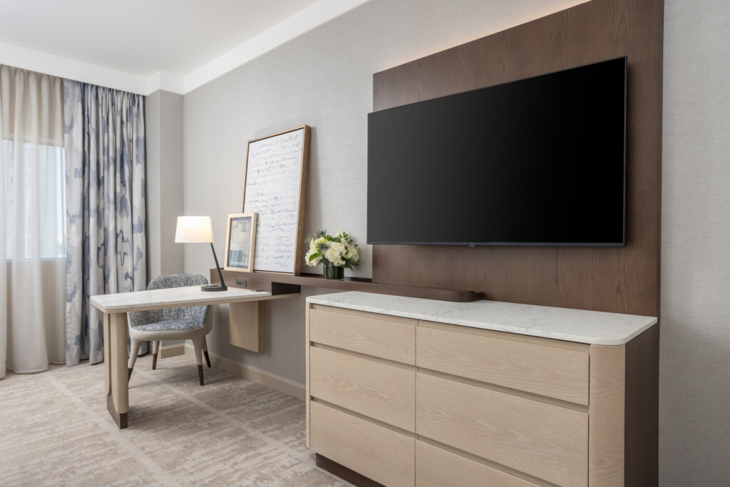 Modern hotel guest room with light wood dresser, wall-mounted television, work desk, and sitting area with patterned curtains