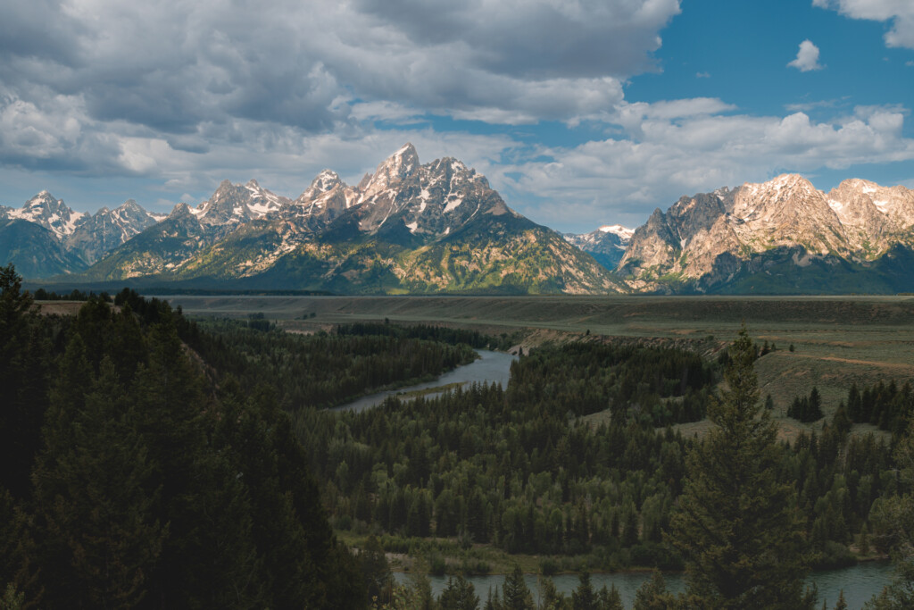 Snow-capped mountain peaks rise behind a forested valley with a winding river and meadows under blue sky with clouds
