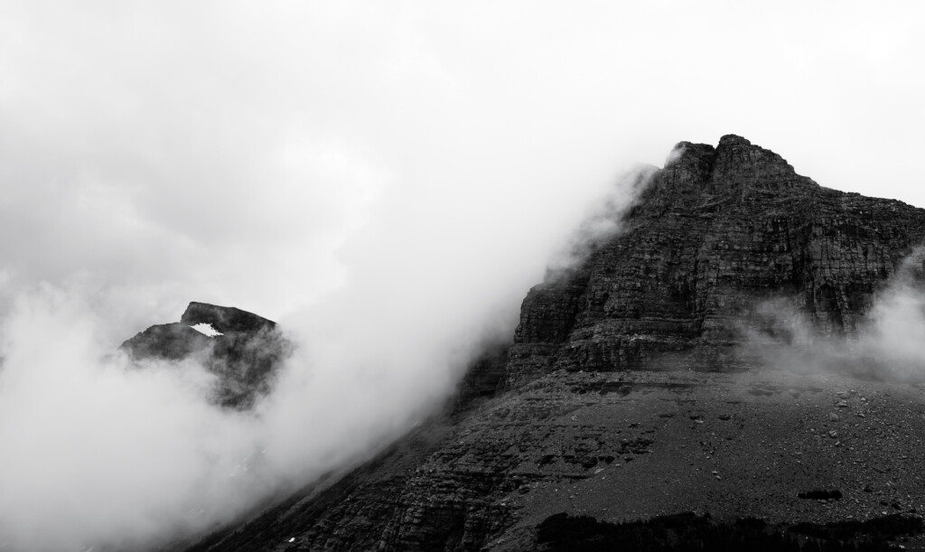 Rugged mountain peaks emerging from thick fog and low clouds, layered rock formations with sparse vegetation visible on steep slopes.