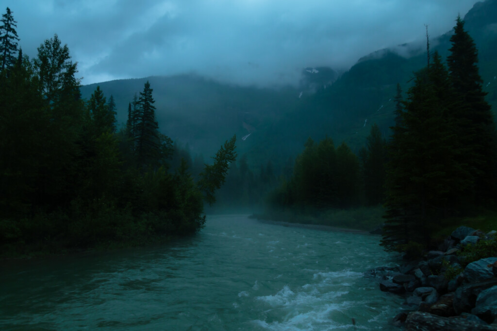 Turquoise river flows through forested mountain valley under stormy sky, evergreen trees lining both banks with misty peaks beyond.