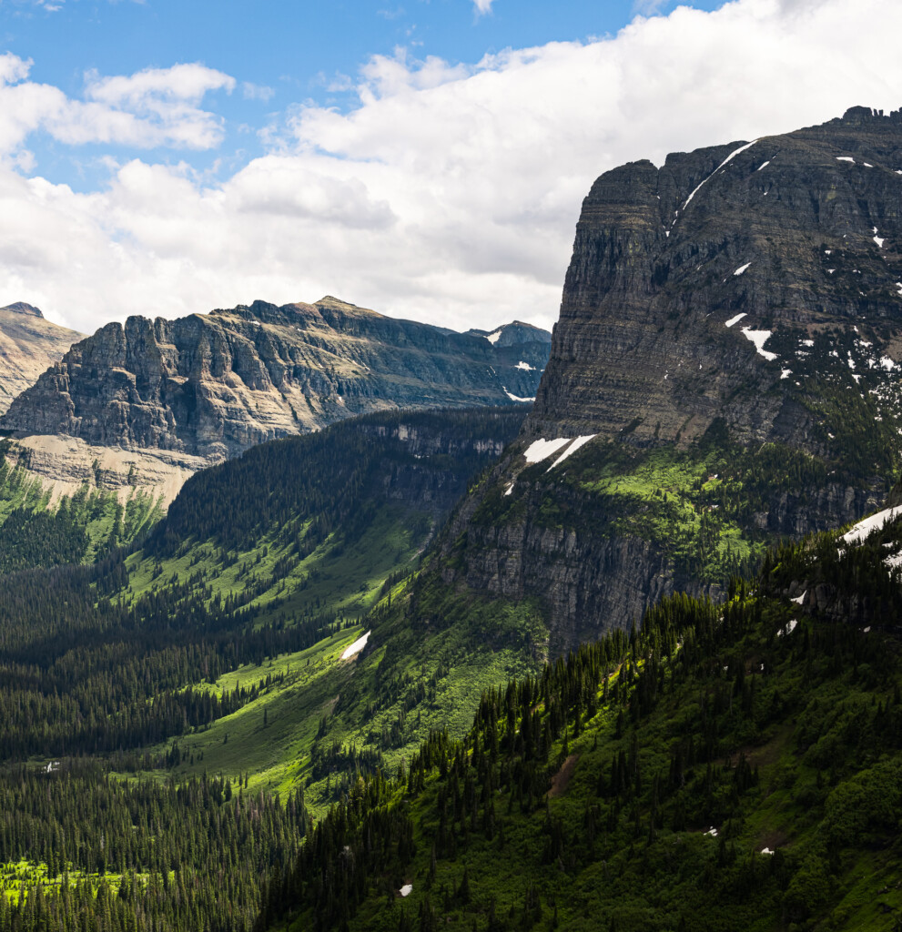 Dramatic mountain valley with steep layered rock cliffs, evergreen forest covering slopes, and patches of snow dotting the peaks under a blue sky with white clouds