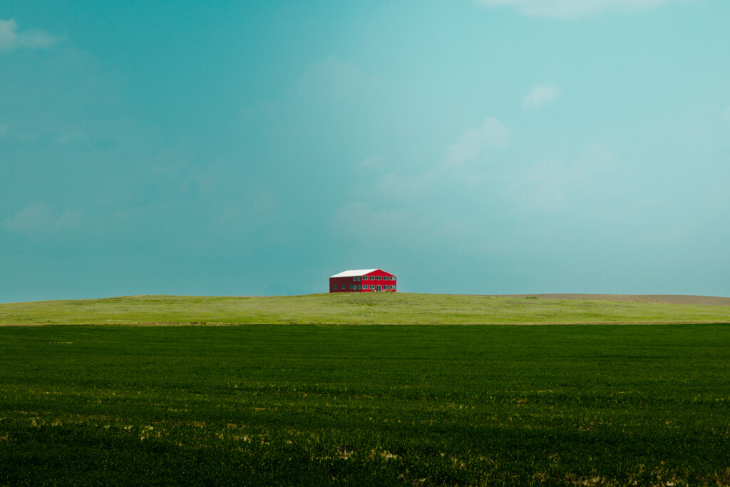 A red barn with white roof sits alone on a grassy hilltop under a clear turquoise sky