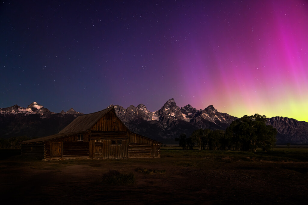 A weathered log cabin stands in an open field beneath a dramatic purple and pink aurora, jagged mountain peaks silhouetted in the background.
