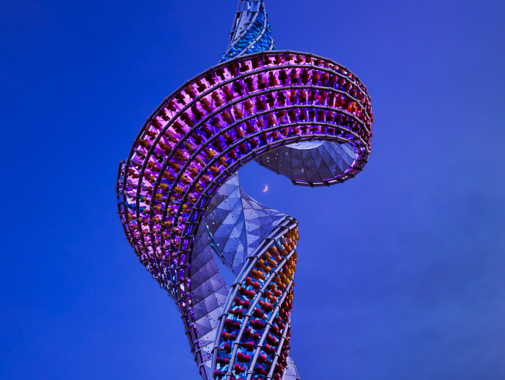 Illuminated spiral tower structure at dusk with purple and red lighting against deep blue sky