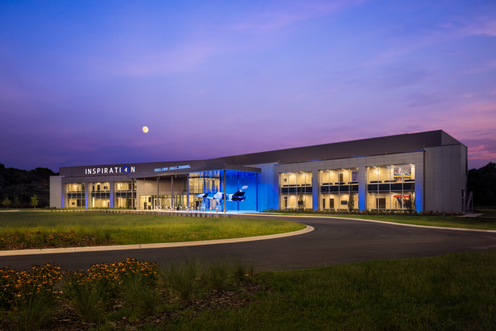 Modern institutional building illuminated at dusk with blue accent lighting, manicured landscaping and curved driveway in foreground