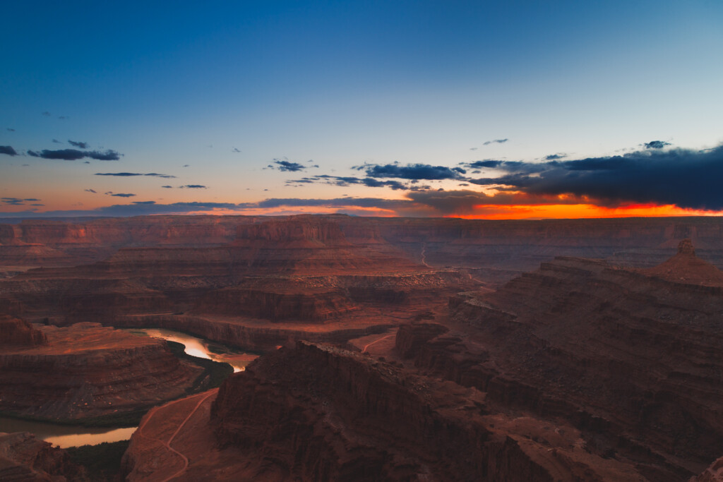 Dramatic sunset illuminating layered canyon walls and a winding river below, with orange sky and cloud formations above the vast desert terrain.